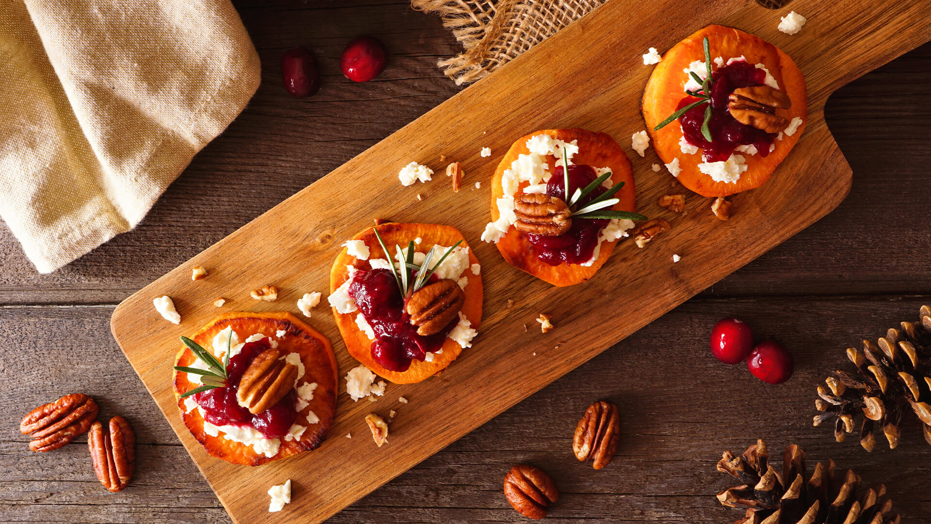 Festive sweet potato rounds topped with pecans, cranberry, rosemary, and goat chesse sit on a wood cutting board.